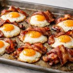Eggs baked on a sheet pan alongside bacon, creating a simple combination of set whites, soft yolks, and crisp strips in one tray.
